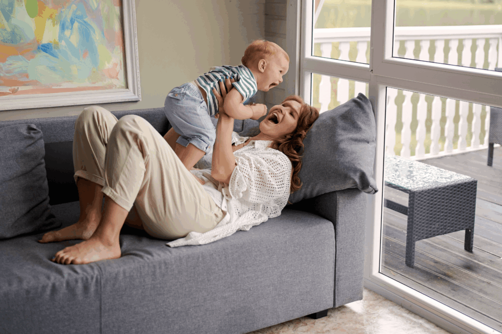 A woman lies on a couch, smiling as she lifts a laughing baby near a window.